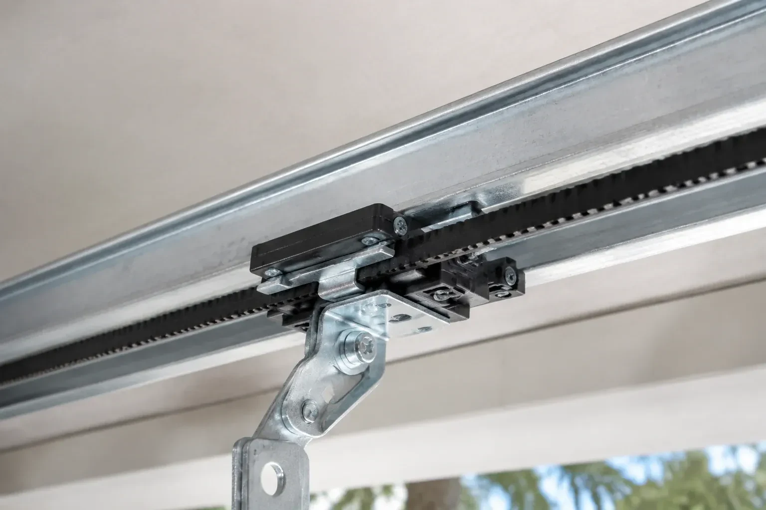 Black plastic trolley and bent galvanized bracket on a steel opener rail with a toothed black belt, against a ceiling. — Belt-Drive Opener Repair, Markham