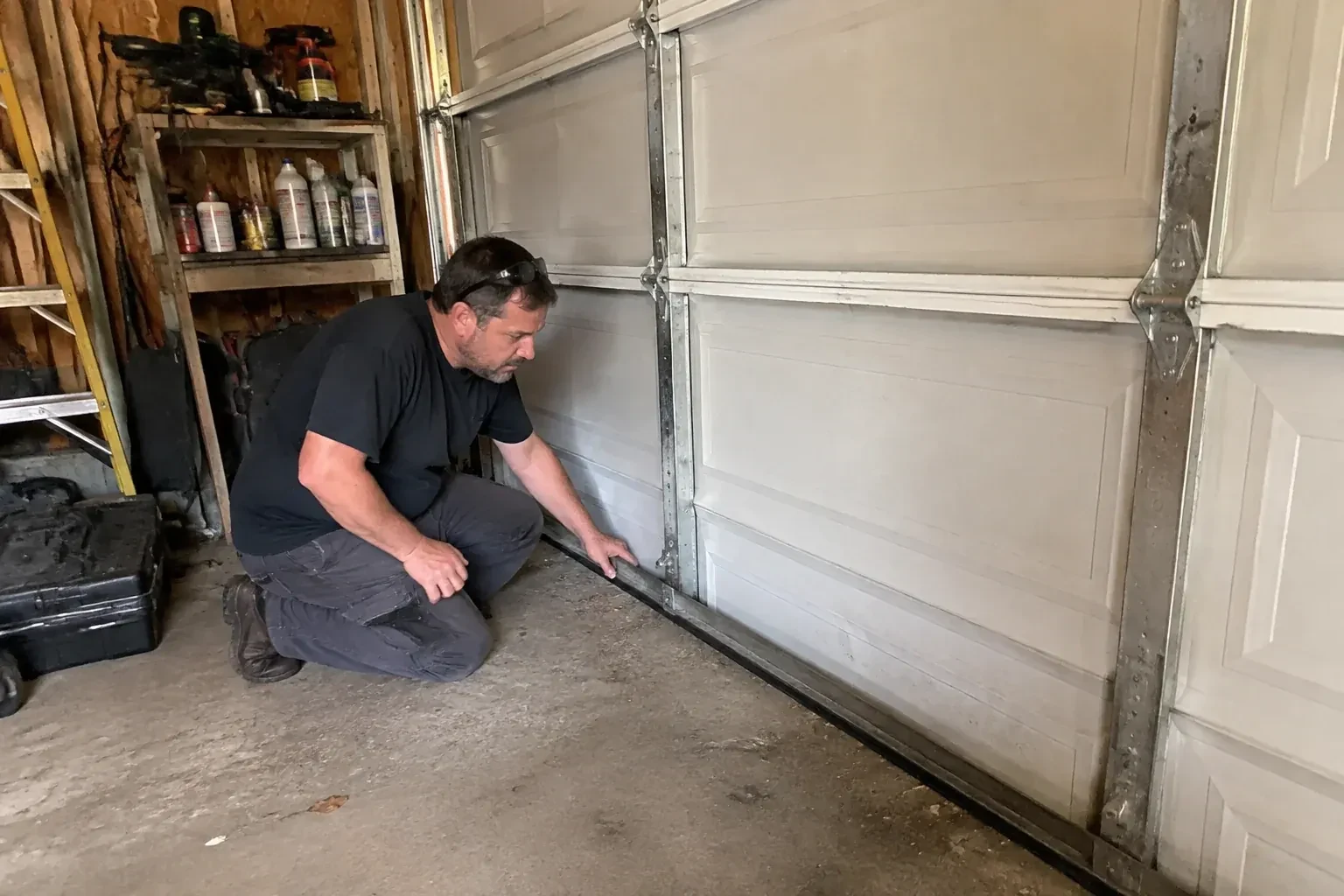 Person kneeling by a white paneled door in a garage, one hand on the black seal along its bottom edge. — Garage Door Roller Replacement, Markham