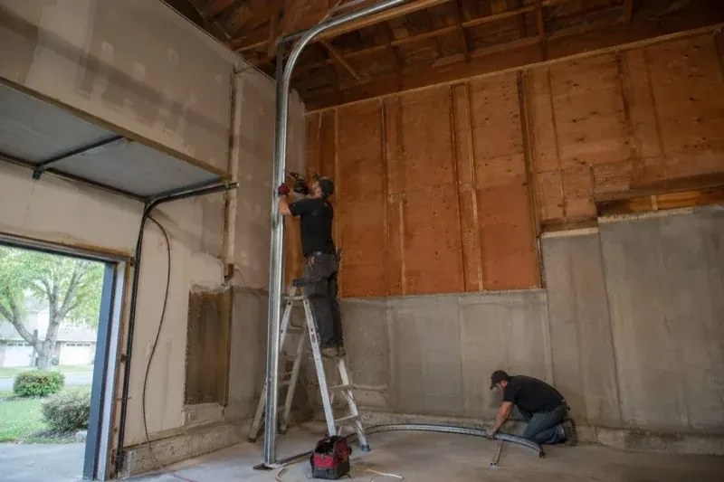 One person on a stepladder working beside a tall curved garage-door track while another kneels beside loose track sections in an unfinished garage. — High-Lift Garage Door Conversion, Markham