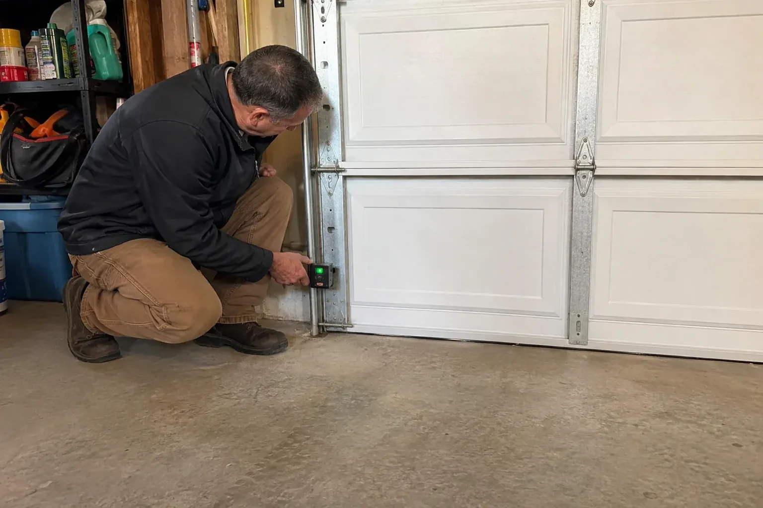 Technician kneeling beside a white paneled garage door, holding a small black box with a green light against the vertical track. — Photo-Eye Sensor Alignment or Replacement, Markham